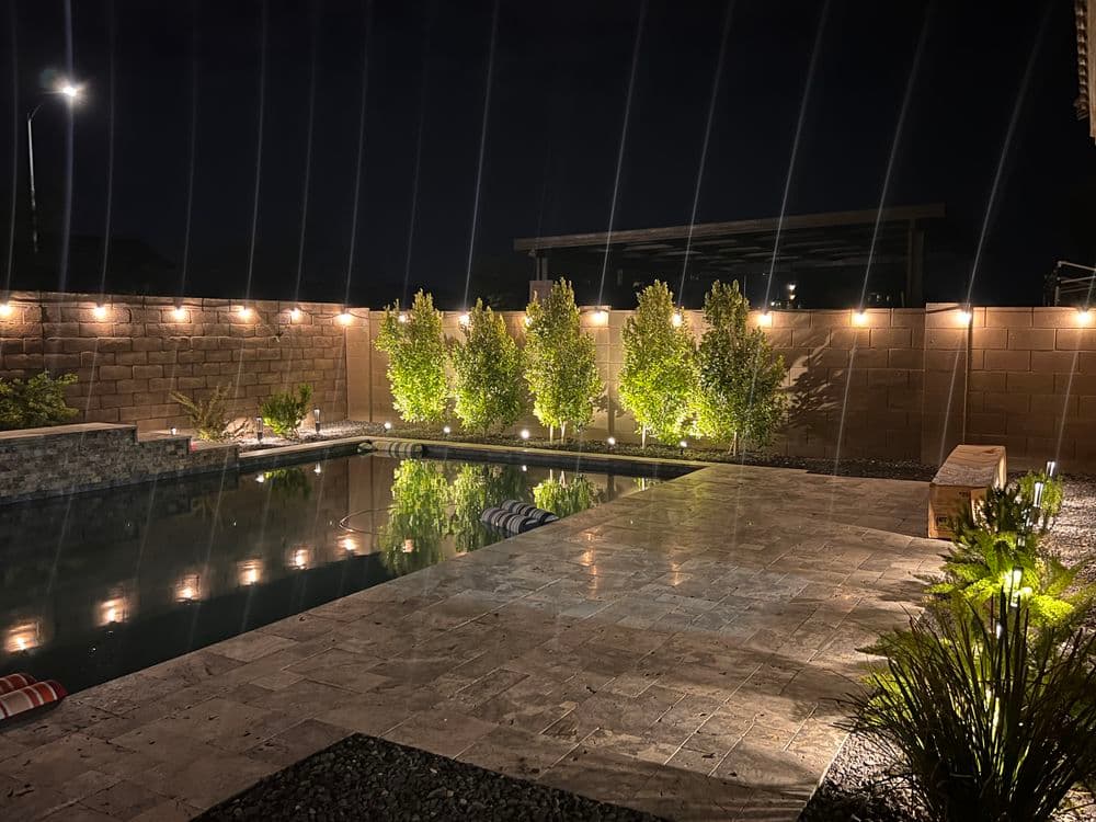Nighttime backyard pool with illuminated greenery and stone patio, creating a serene ambiance.