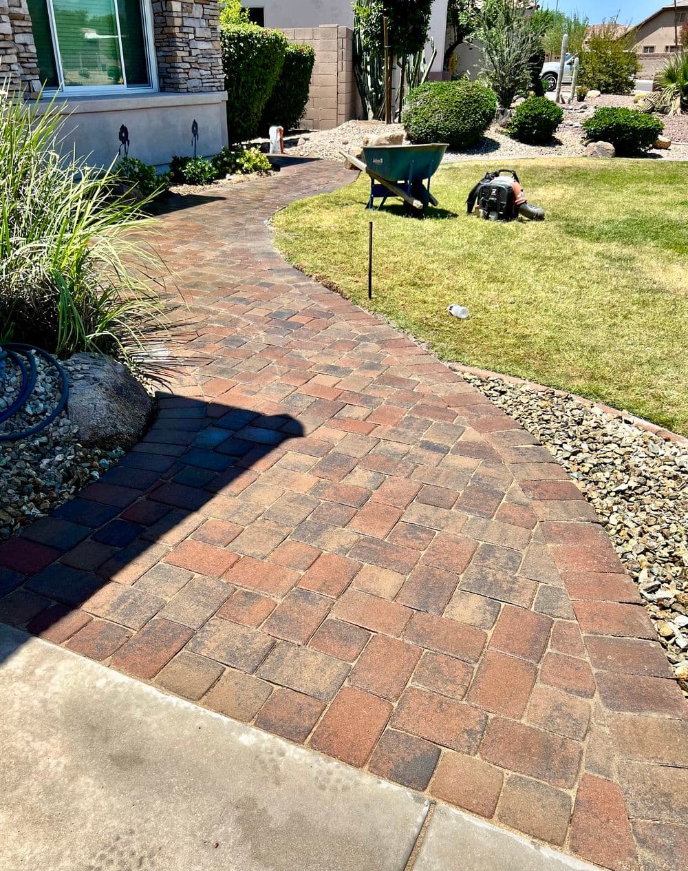 Paved pathway winding through a landscaped garden with wheelbarrow and grass lawn.
